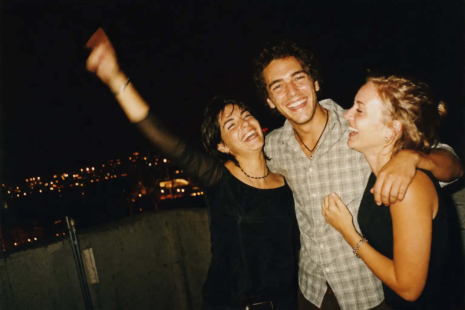 Three friends laughing on a rooftop at night, lit by harsh direct flash against a dark city background